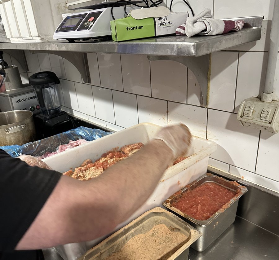 Yassas chef layering dry herb blend including imported Greek oregano onto raw chicken in commercial kitchen tray - 24-hour marination process